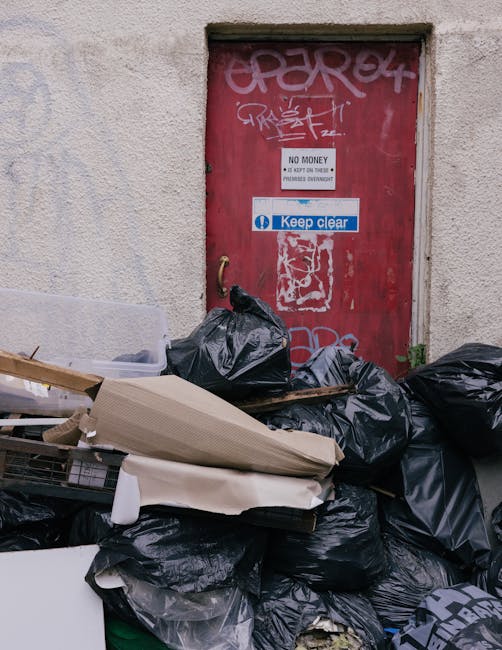 A pile of black rubbish bags, cardboard boxes, and discarded packaging materials are stacked in front of a red metal door, which is covered in graffiti and stickers. The bags appear full and are place
