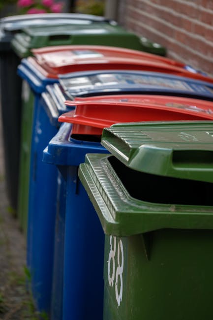 A green, outdoor waste bin with a semi-transparent lid containing partially visible collected rubbish, including crumpled paper, plastic packaging, and other mixed waste items. The bin features a circ